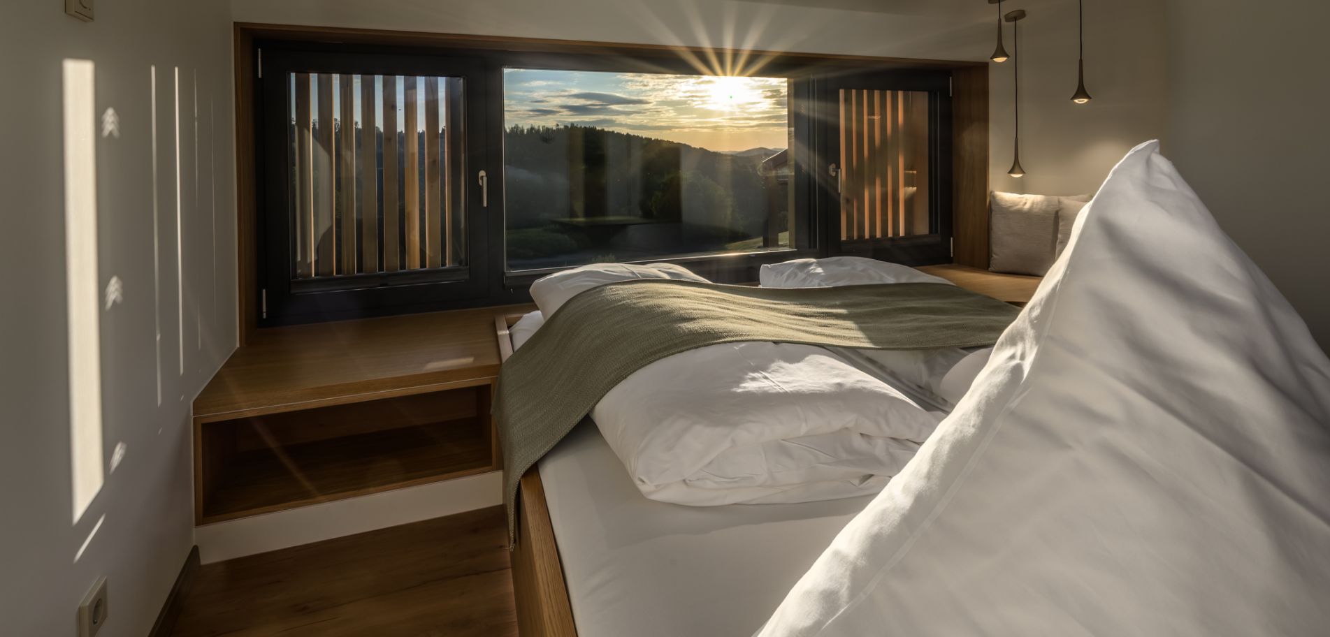 Schlafzimmer im Refugium siehm mit Doppelbett, Eckfensterfront und tief stehender Sonne über den bewaldeten Hügeln